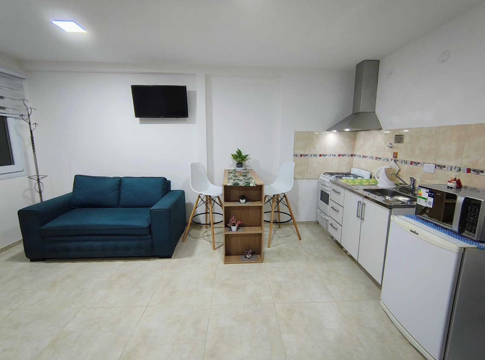 A compact living space with a sofa, a small dining table with chairs, a kitchen area with a stove and microwave, and a flat-screen TV mounted on the wall.
