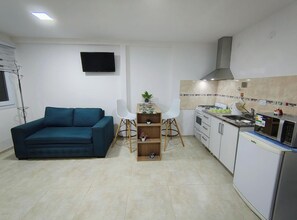 Espaço compacto com sofá, pequena mesa de jantar com cadeiras, cozinha equipada com fogão e micro-ondas, e televisão de tela plana na parede.