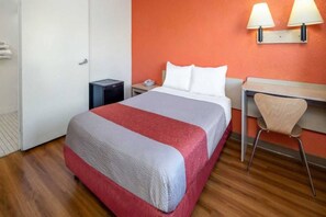 A hotel room with a bed, a desk, a chair, a nightstand, and a lamp.