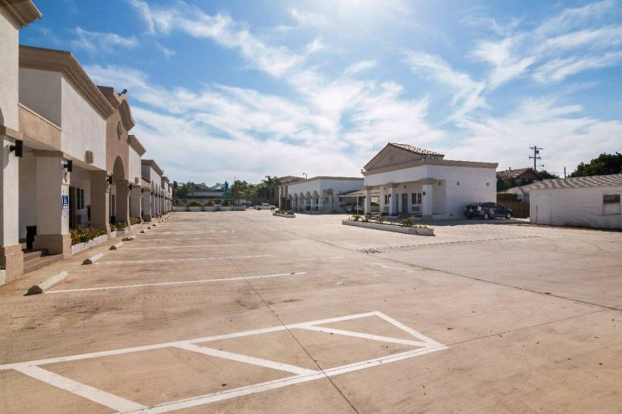A parking lot with multiple buildings, including a large white structure with a flat roof and smaller buildings with arched windows.