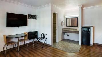 A room with a flat-screen TV mounted on the wall, a small table with chairs, a compact refrigerator, and a bathroom with a sink and mirror.