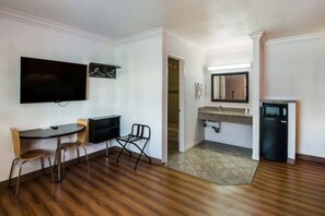 Una habitación con una televisión de pantalla plana montada en la pared, una mesa pequeña con sillas, un refrigerador compacto y un baño con lavamanos y espejo.