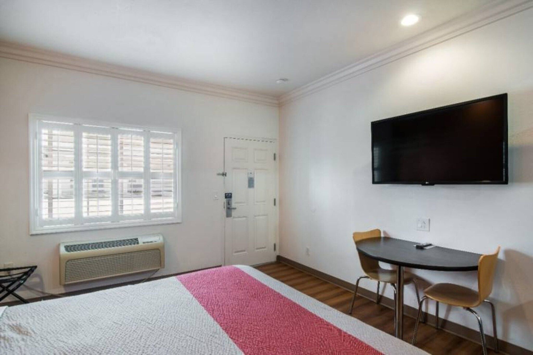 A room with a bed, a desk, two chairs, a television, a window with blinds, and a door.