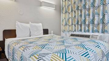 Une chambre d’hôtel avec un lit, des oreillers blancs, une couvre-lit à motifs, un climatiseur fixé au mur et un rideau à motifs géométriques.