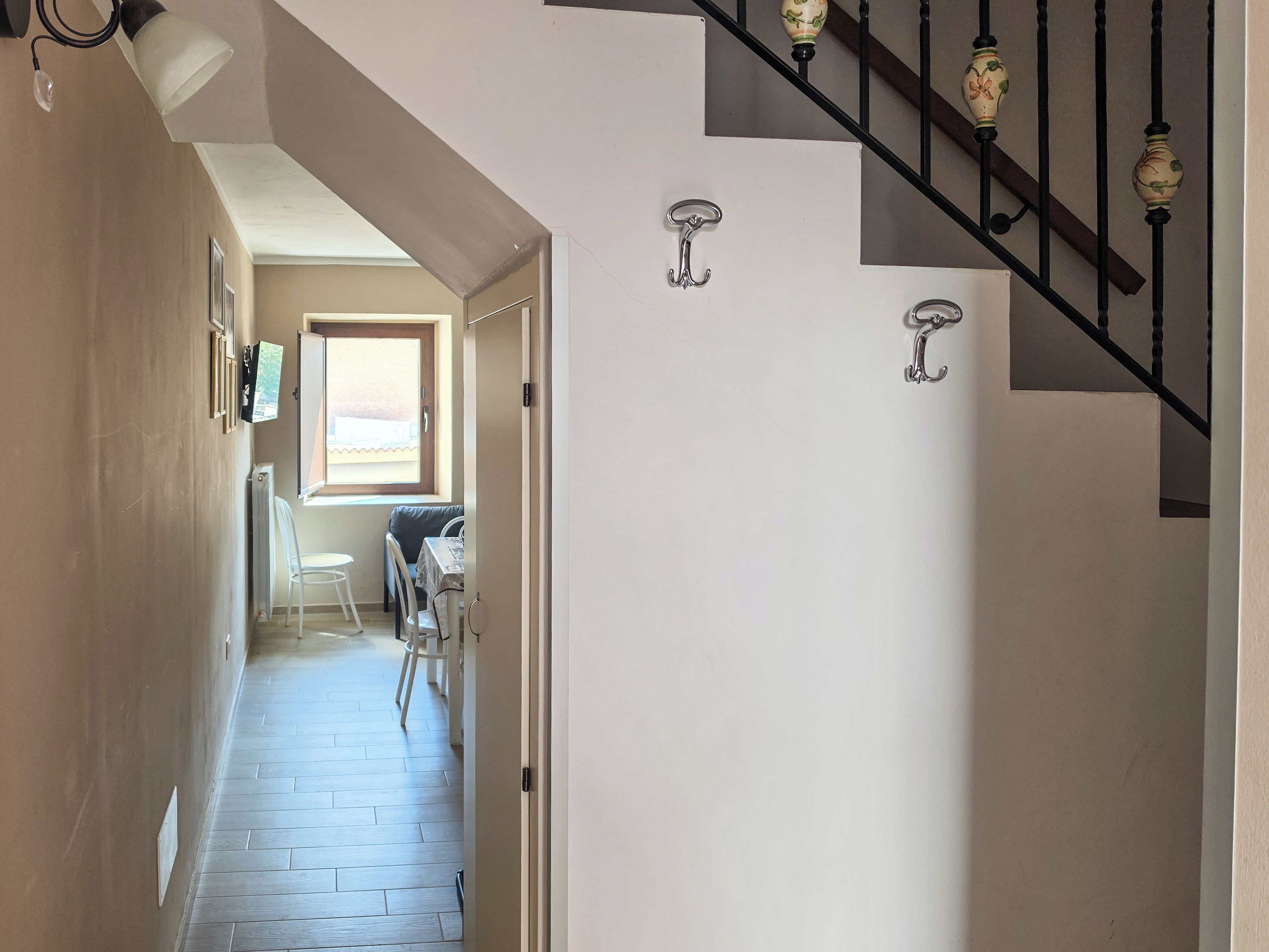 Un couloir mène à une salle à manger équipée d’une table et de chaises, d’une fenêtre avec des stores et d’un escalier agrémenté d’éléments décoratifs.