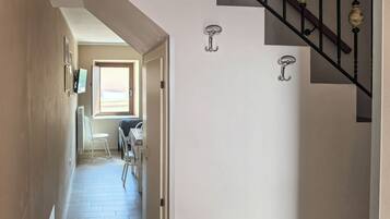 Un couloir mène à une salle à manger équipée d’une table et de chaises, d’une fenêtre avec des stores et d’un escalier agrémenté d’éléments décoratifs.