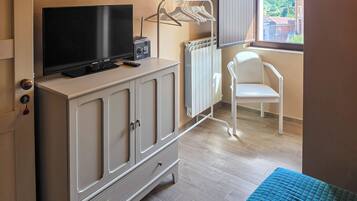Une pièce avec un téléviseur posé sur un meuble, un radiateur, une chaise blanche et une fenêtre donnant sur des immeubles.