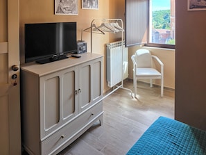 Une pièce avec un téléviseur posé sur un meuble, un radiateur, une chaise blanche et une fenêtre donnant sur des immeubles.