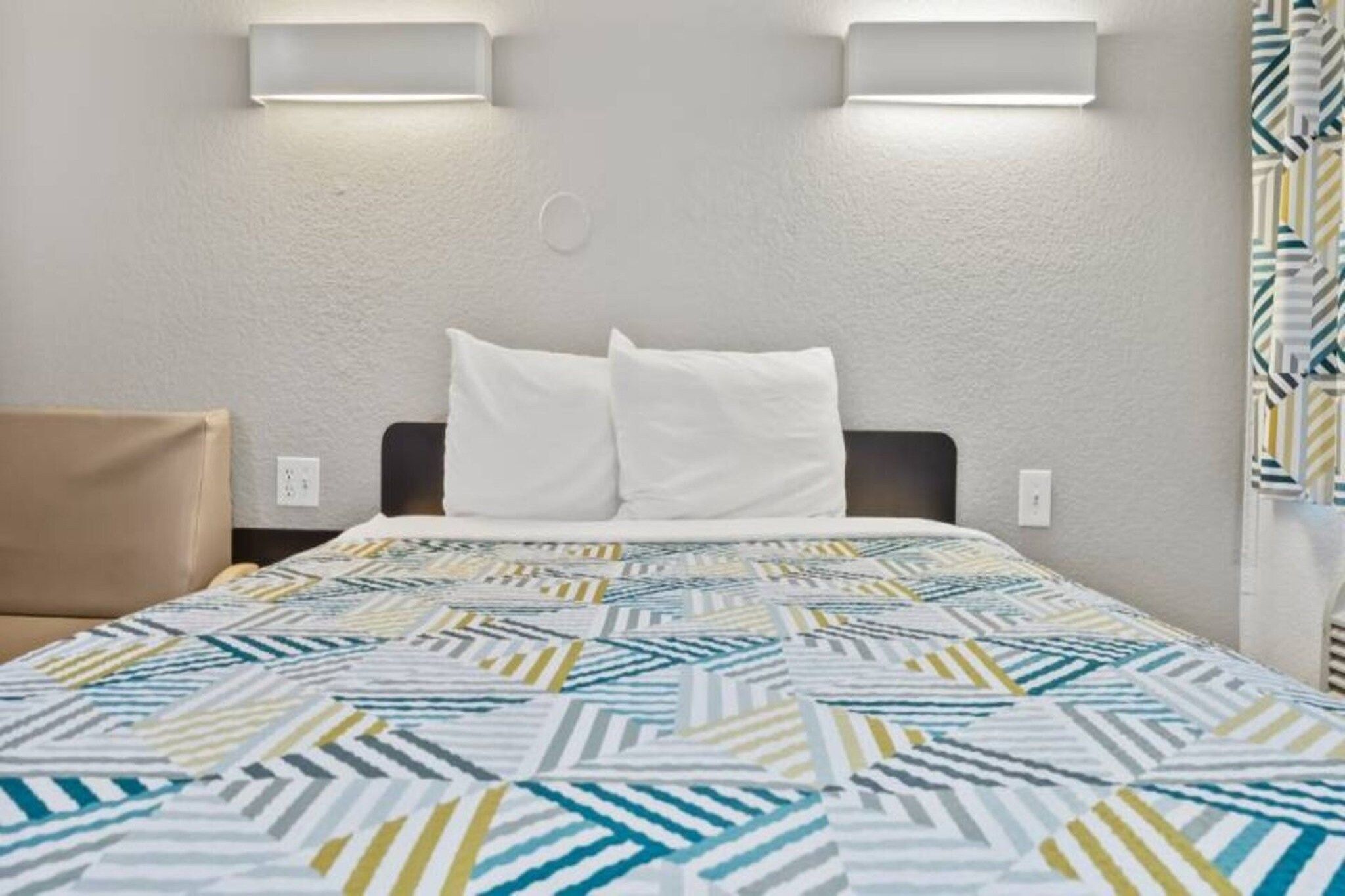 Habitación de hotel con cama, dos almohadas, colcha estampada, cabecero e iluminación en la pared.