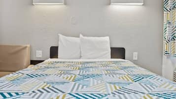 Habitación de hotel con cama, dos almohadas, colcha estampada, cabecero e iluminación en la pared.