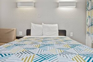 Habitación de hotel con cama, dos almohadas, colcha estampada, cabecero e iluminación en la pared.