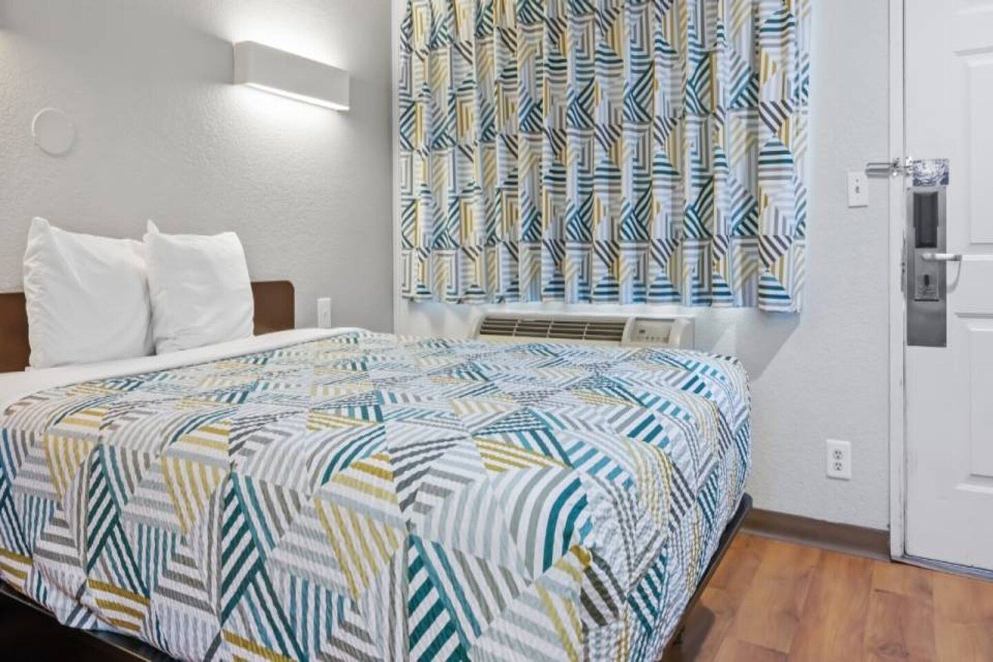 A hotel room with a bed, a wall-mounted air conditioner, and a door.