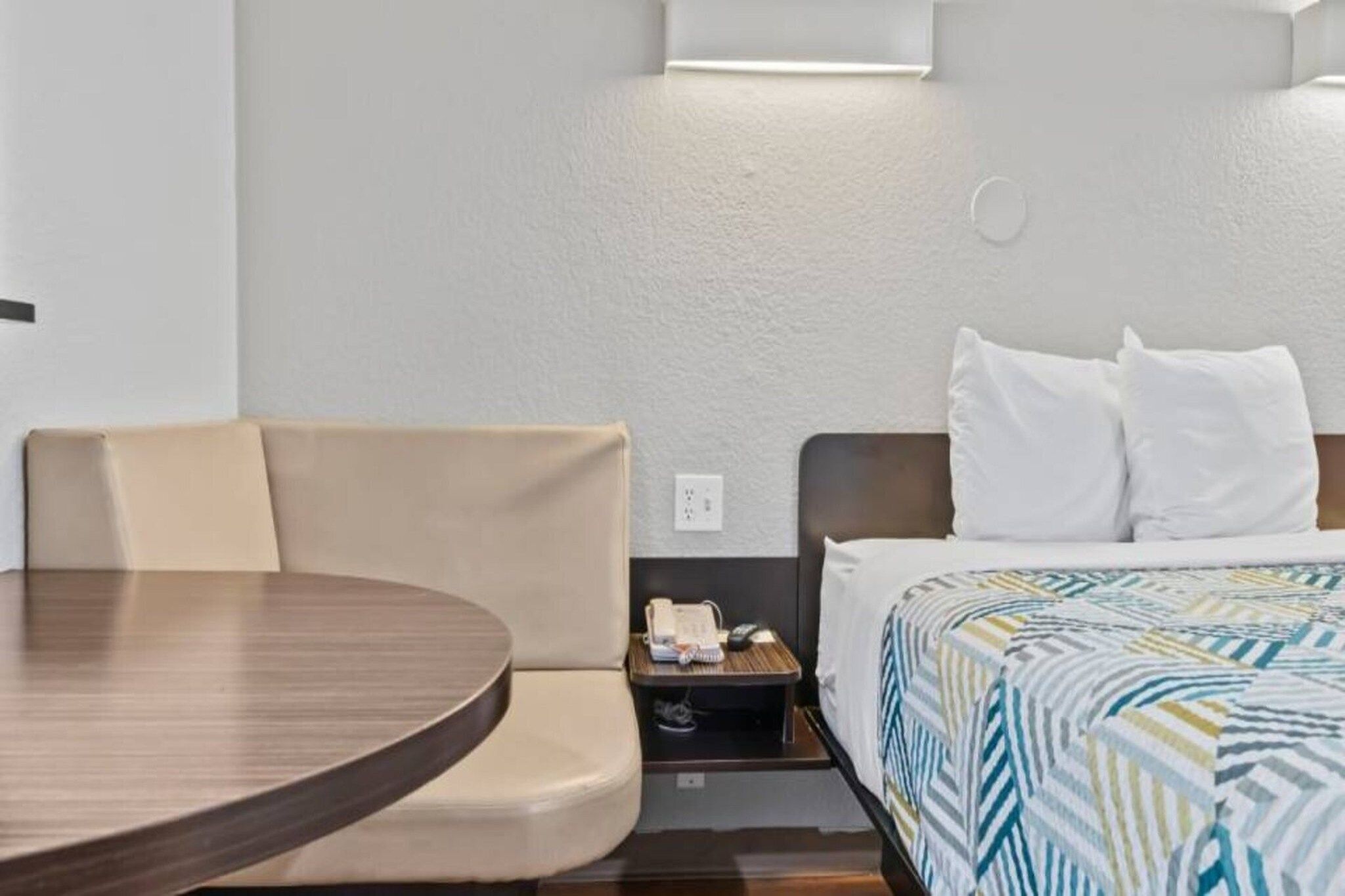 A hotel room with a bed, a small bedside table with a phone, a chair, and a round table.