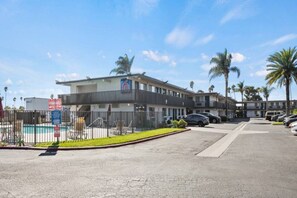 Un hotel con zona de piscina vallada, varios espacios de estacionamiento y cielo despejado.