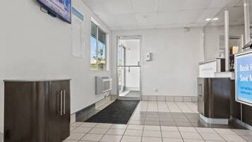A hotel lobby with a reception desk, a digital screen, a door leading to a balcony, and a tiled floor.