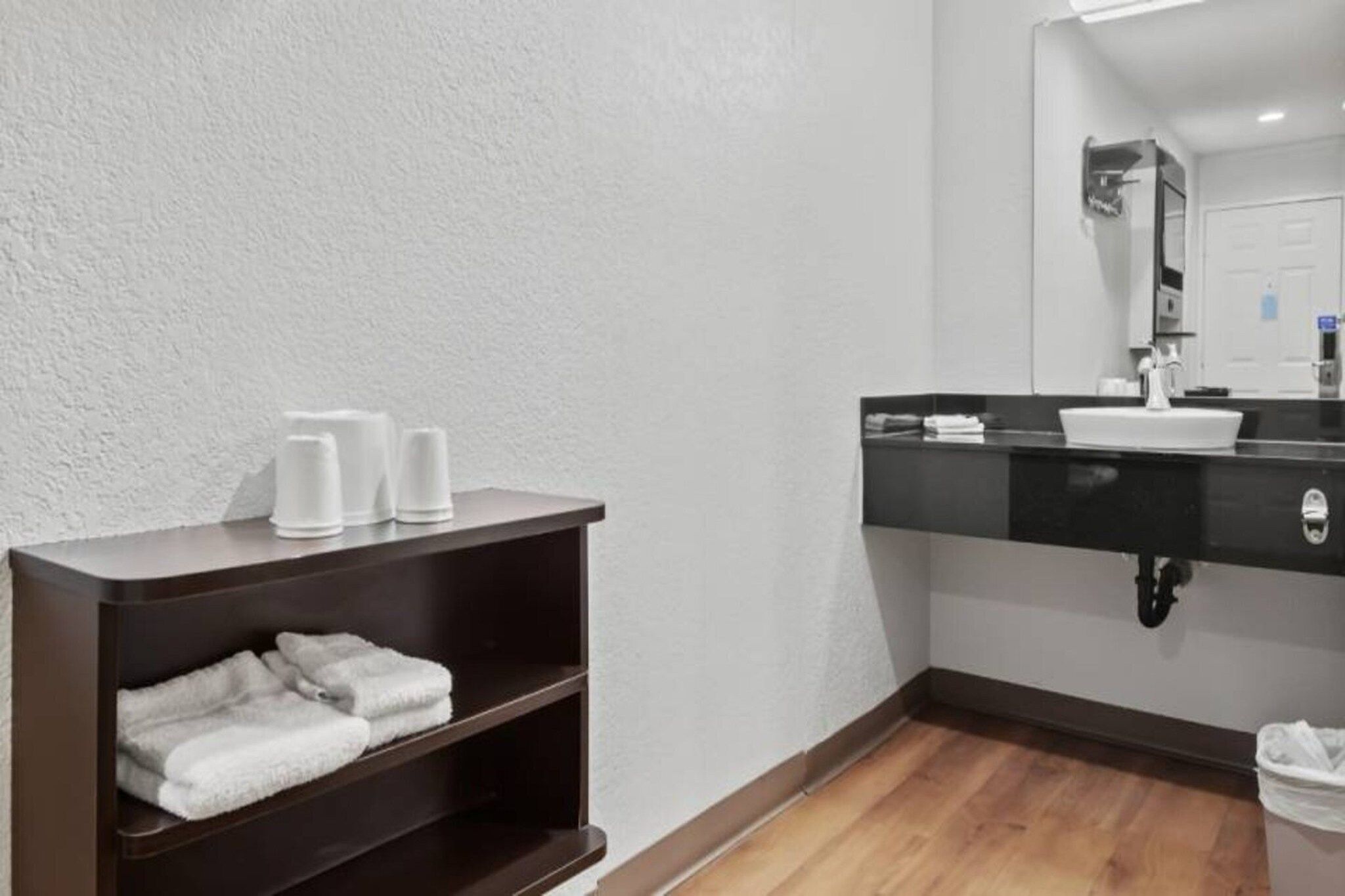 A bathroom with a dark wooden shelf holding towels, a white sink with a black countertop, and a mirror above it.