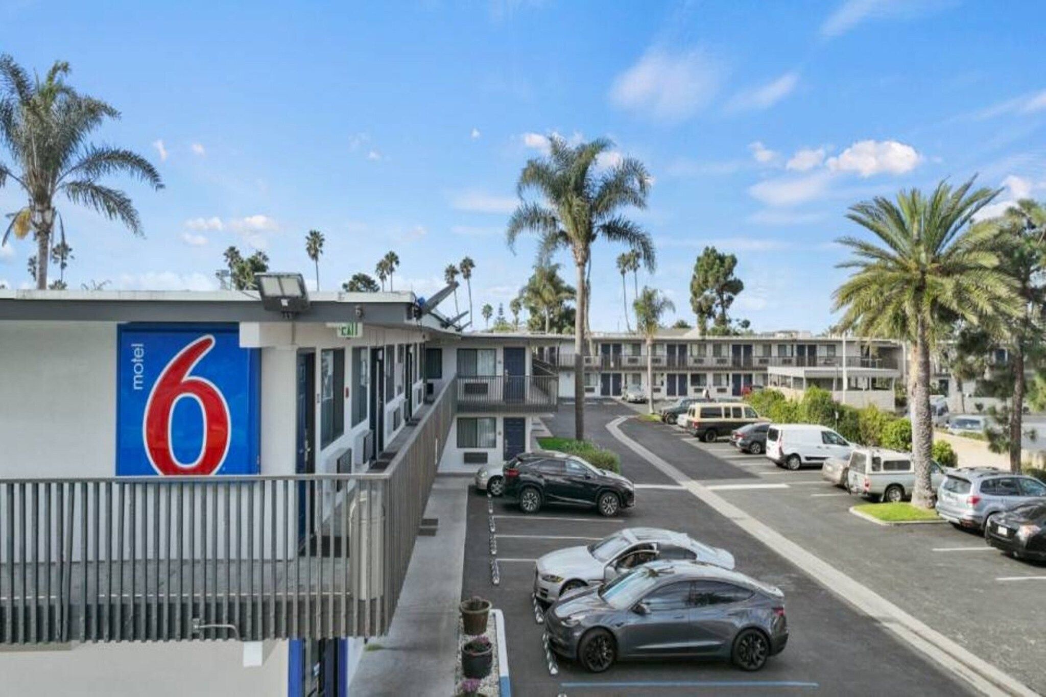 A motel with a prominent number sign, a parking lot with cars, and palm trees in the background.