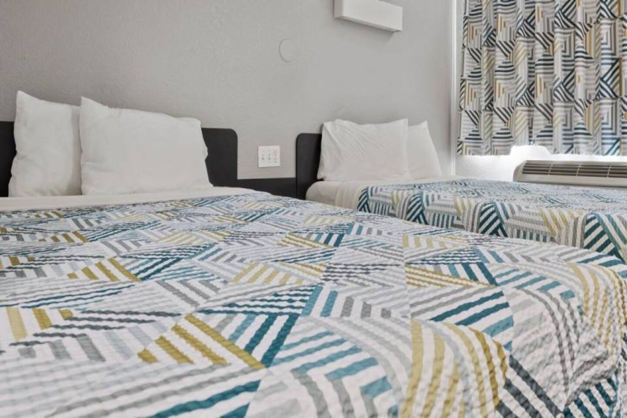 A hotel room with two beds, a grey wall, a window with patterned curtains, and a wall-mounted air conditioning unit.