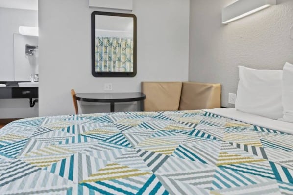 Una habitación de hotel con una cama, una mesita redonda pequeña, una silla, un espejo y una lámpara fijada a la pared.