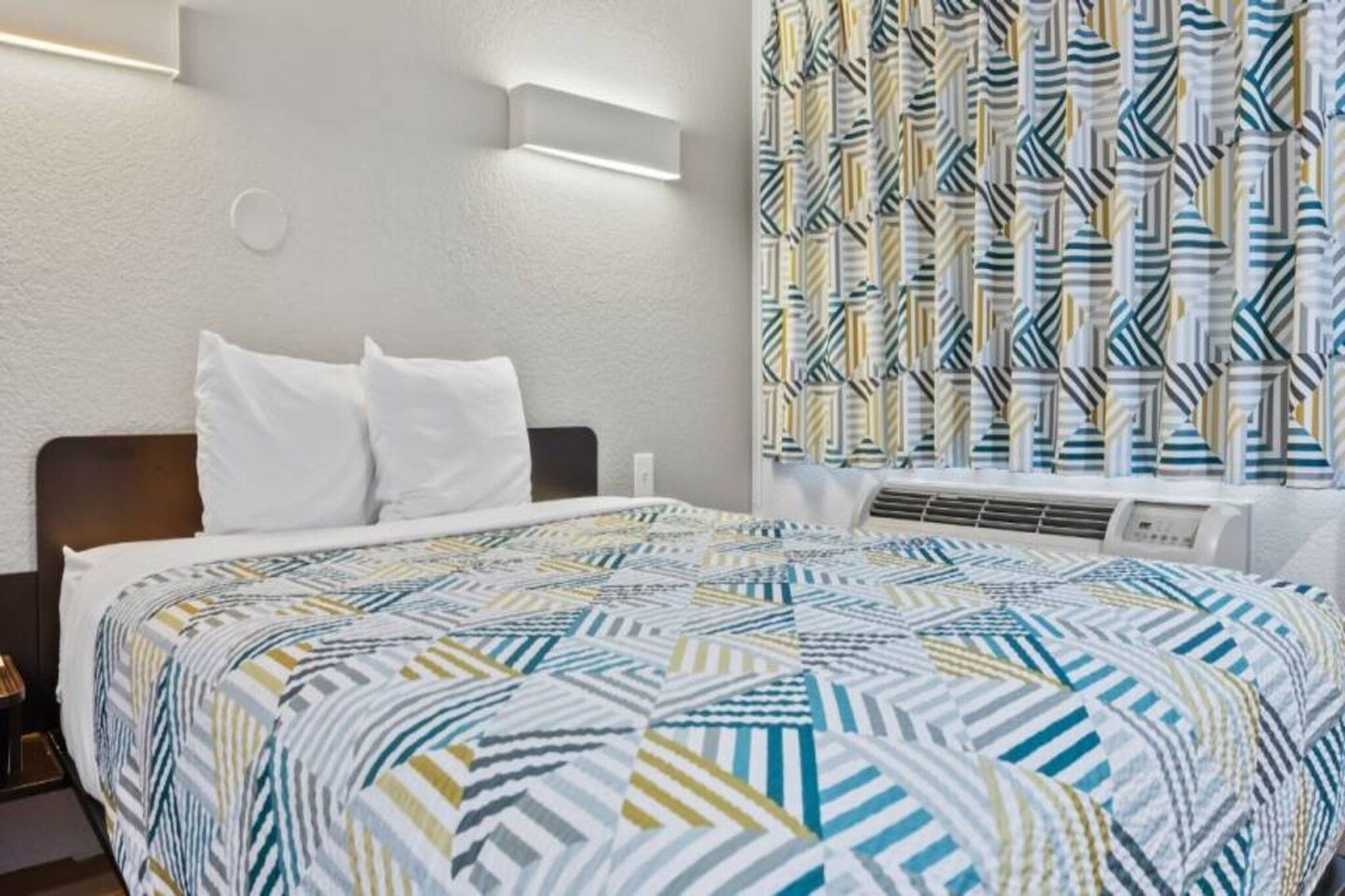 Ett hotellrum med en säng, vita kuddar, ett mönstrat överkast, en väggmonterad luftkonditioneringsanläggning och en gardin med ett geometriskt mönster.