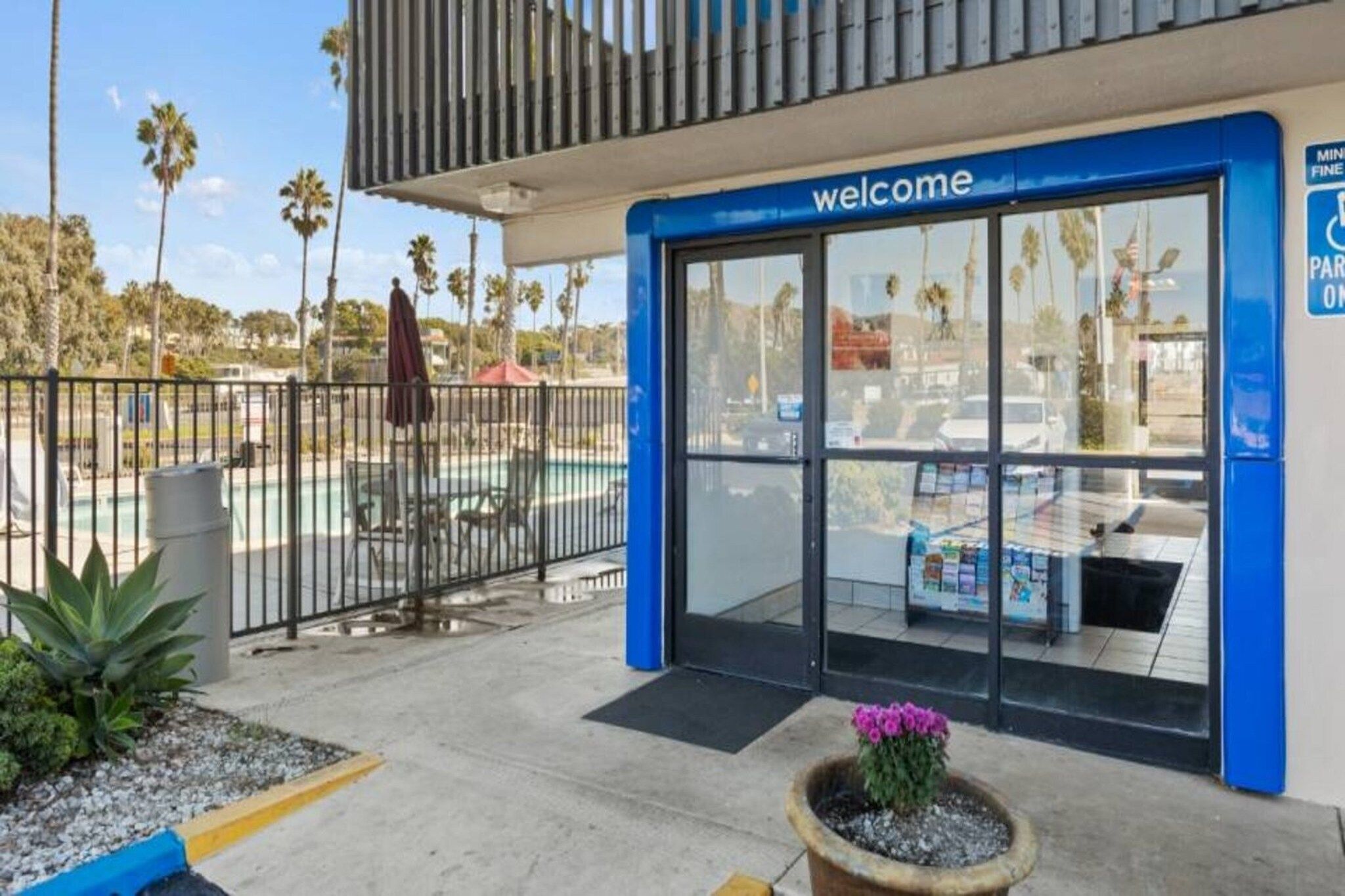 A building entrance with a 'welcome' sign, a gated parking area, and a small garden with a potted plant.