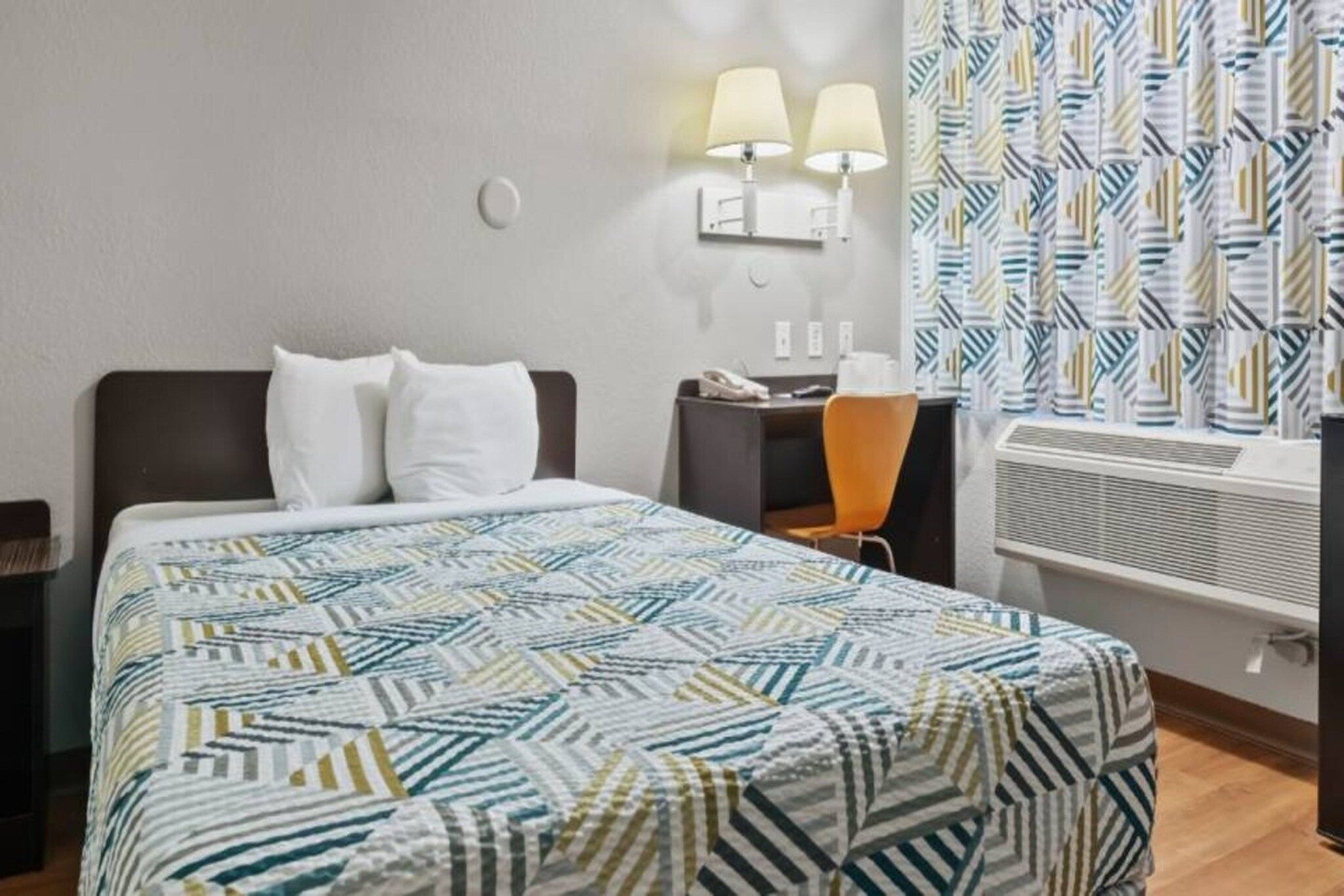 A hotel room with a bed, two bedside lamps, a desk with a chair, and a window with patterned curtains.