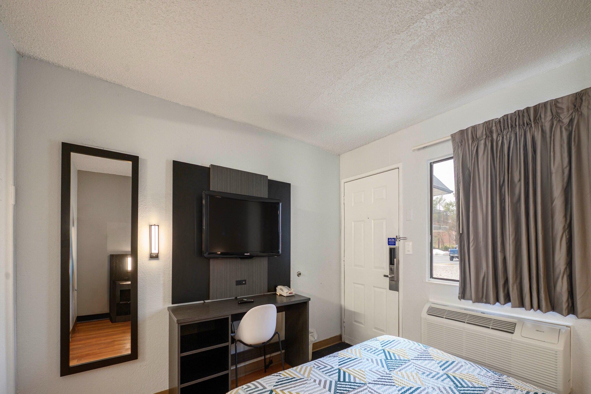 Une chambre d’hôtel avec un lit, un bureau, une chaise, une télévision, une porte et une fenêtre avec des rideaux.