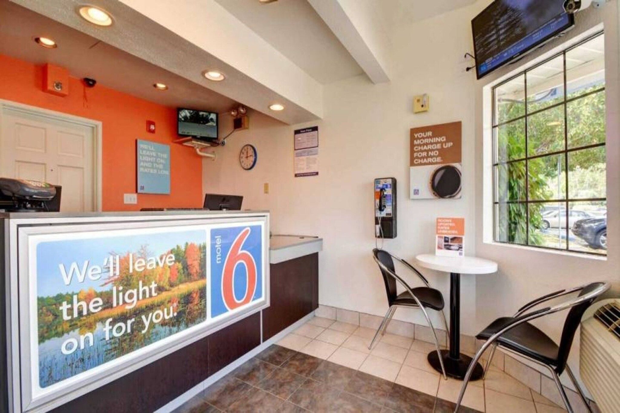 Een hotel receptie met een groot bord waarop een 6-uurs service wordt aangeboden, een klok, een telefoon en een raam met uitzicht op het exterieur.