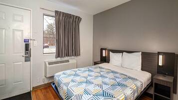 Una habitación de hotel con cama, ventana con cortinas, puerta con cerradura, unidad de aire acondicionado y mesita de noche con lámpara.