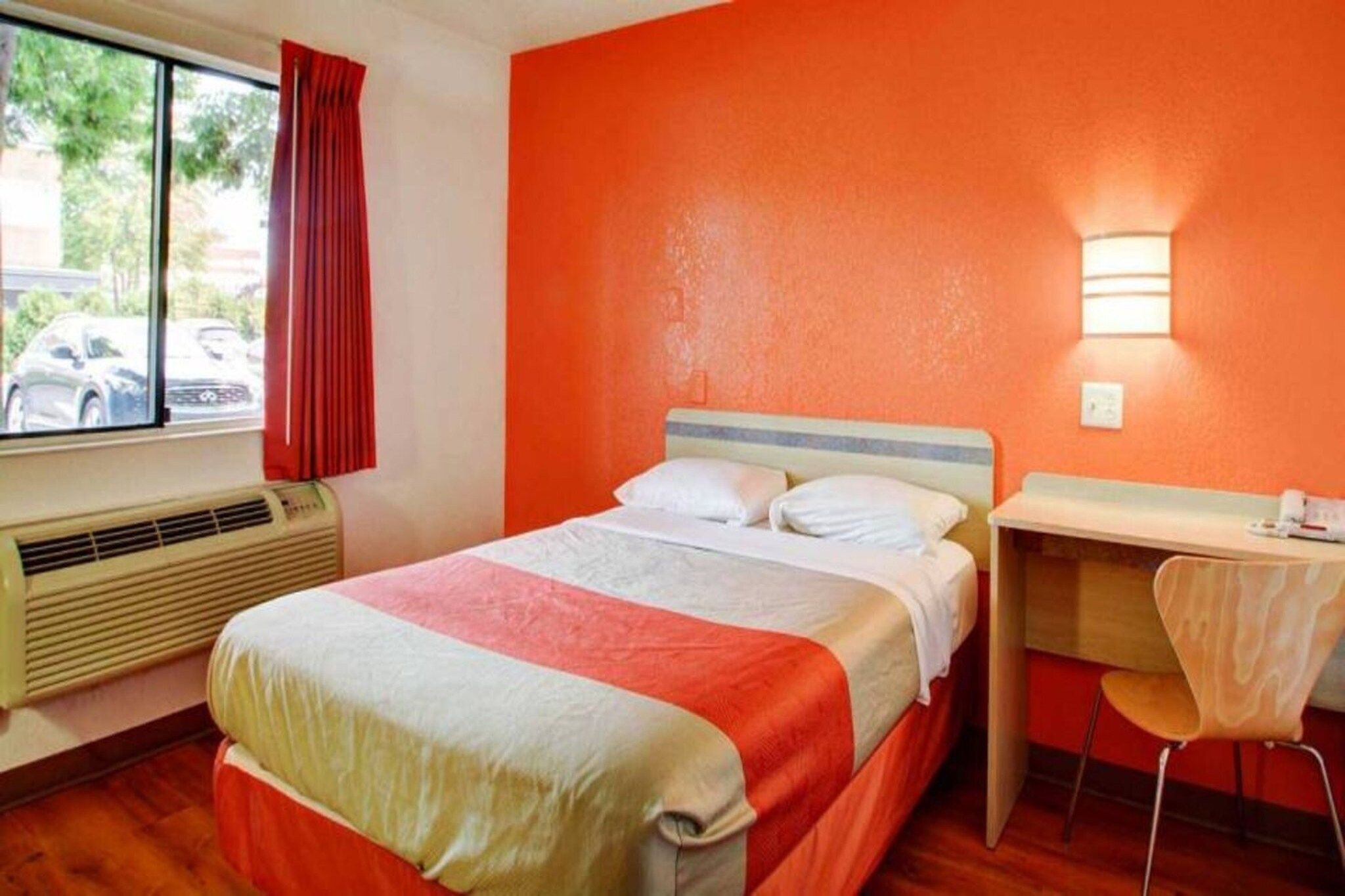 Een hotelkamer met een bed, een bureau en een stoel. Er is een raam met uitzicht op geparkeerde auto's.