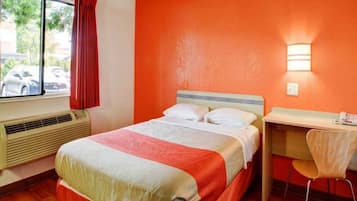 Een hotelkamer met een bed, een bureau en een stoel. Er is een raam met uitzicht op geparkeerde auto's.