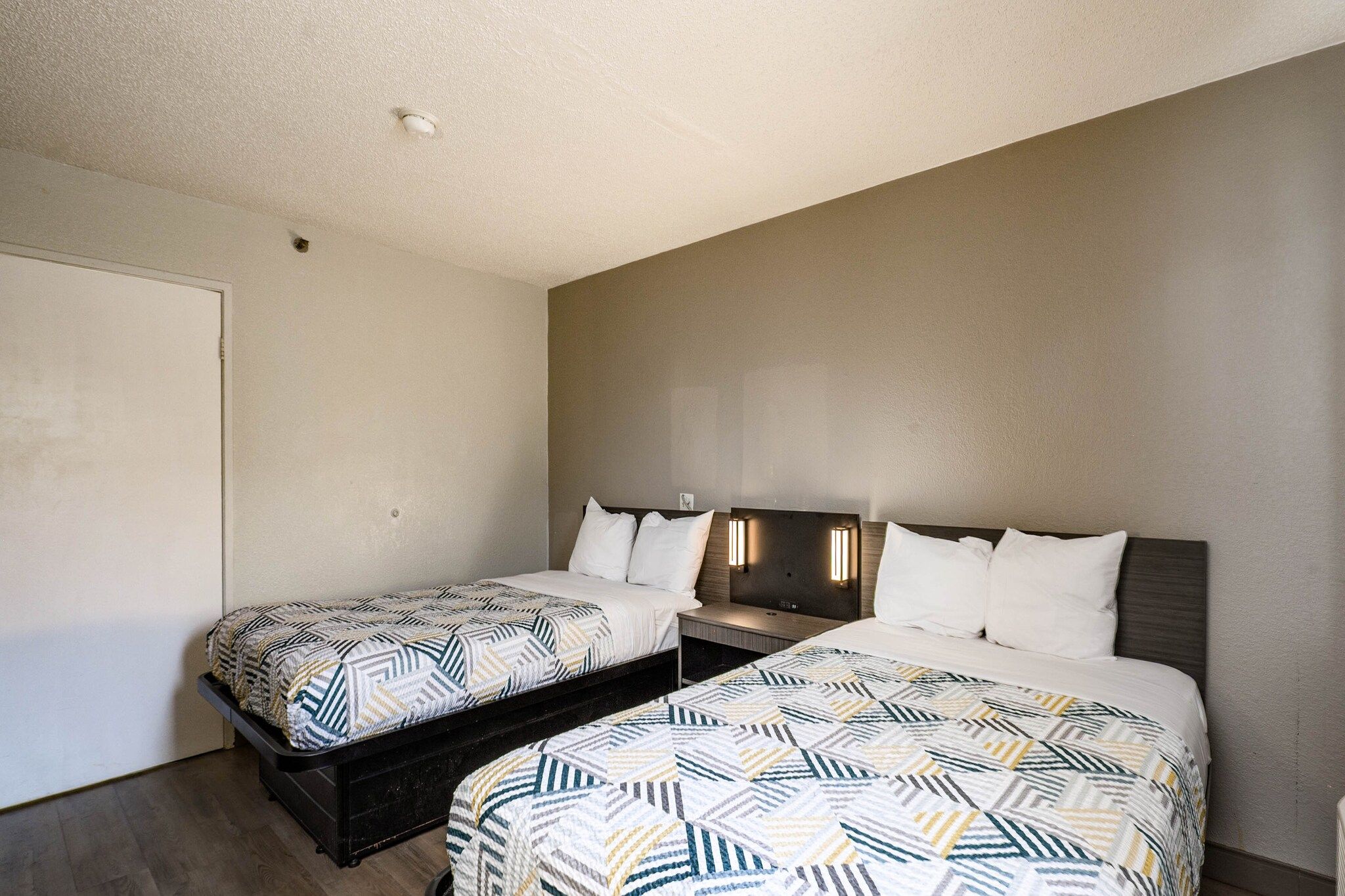 Quarto de hotel com duas camas, uma mesa de cabeceira, uma luminária na parede e uma porta.