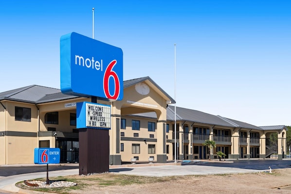 Un motel con un llamativo letrero con el número '6', una entrada despejada y un cartel que anuncia servicios de internet inalámbrico y HBO.