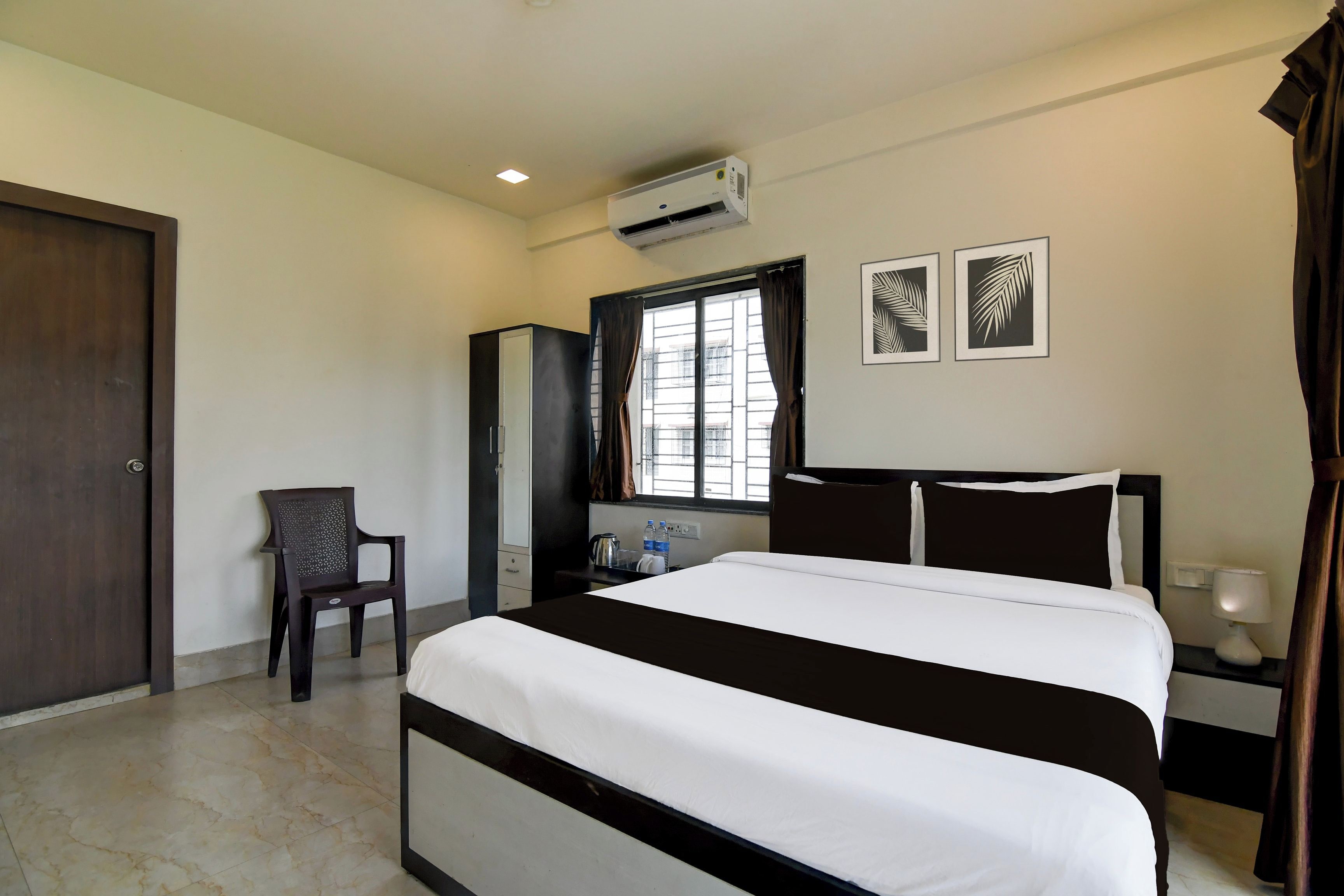 Habitación de hotel con cama, escritorio, silla, equipo de aire acondicionado, ventana con cortinas y dos cuadros en la pared.