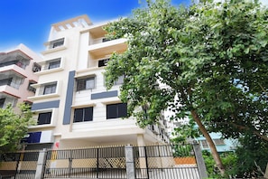 A multi-story residential building with balconies, a gated entrance, and a tree in the foreground.