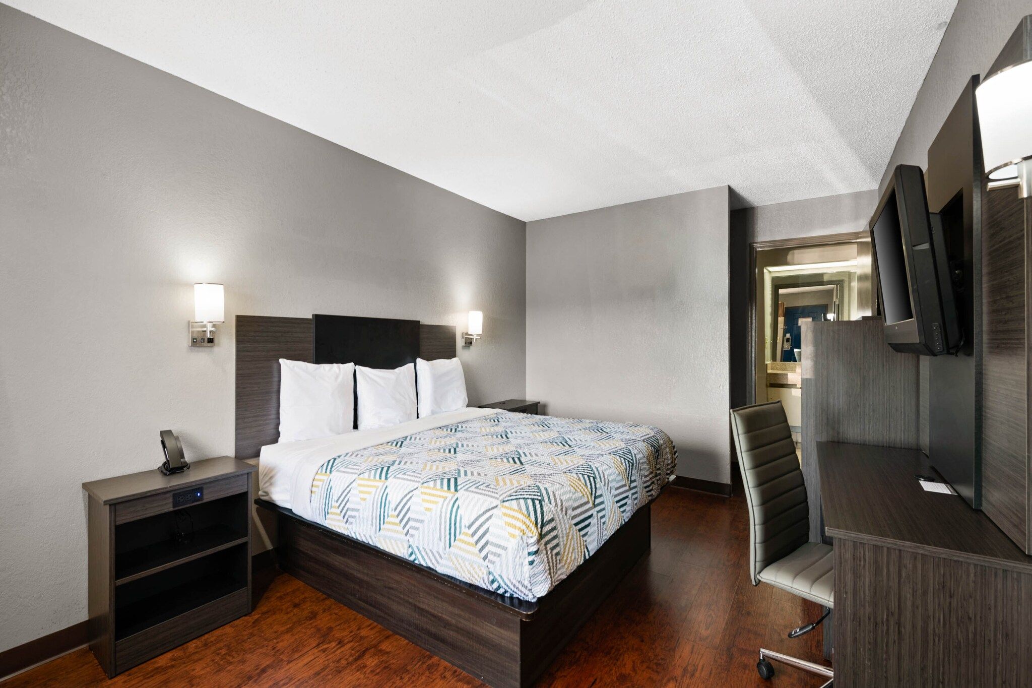 Una habitación de hotel con cama, mesitas de noche, escritorio con silla, televisor y espejo.