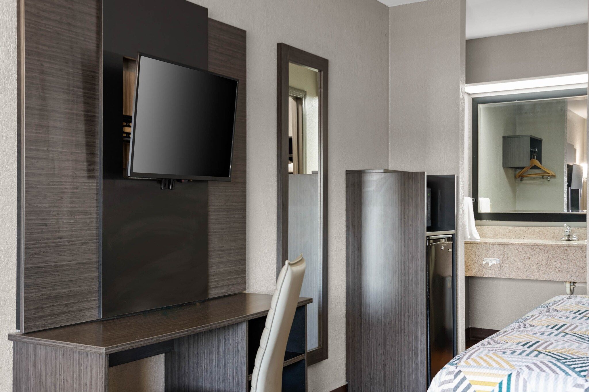 A hotel room with a flat-screen TV mounted on the wall, a mirror, a bed with a patterned bedspread, and a bathroom with a sink and mirror.