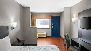 A hotel room with a bed, a desk, a chair, a television, a window with curtains, and a door.