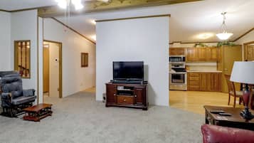 Una sala de estar con televisor, un mueble de entretenimiento de madera, un sofá de cuero rojo, una mesa de comedor de madera y una cocina al fondo.
