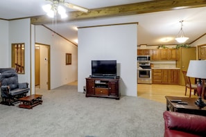 A living room with a television, a wooden entertainment center, a red leather sofa, a wooden dining table, and a kitchen in the background.