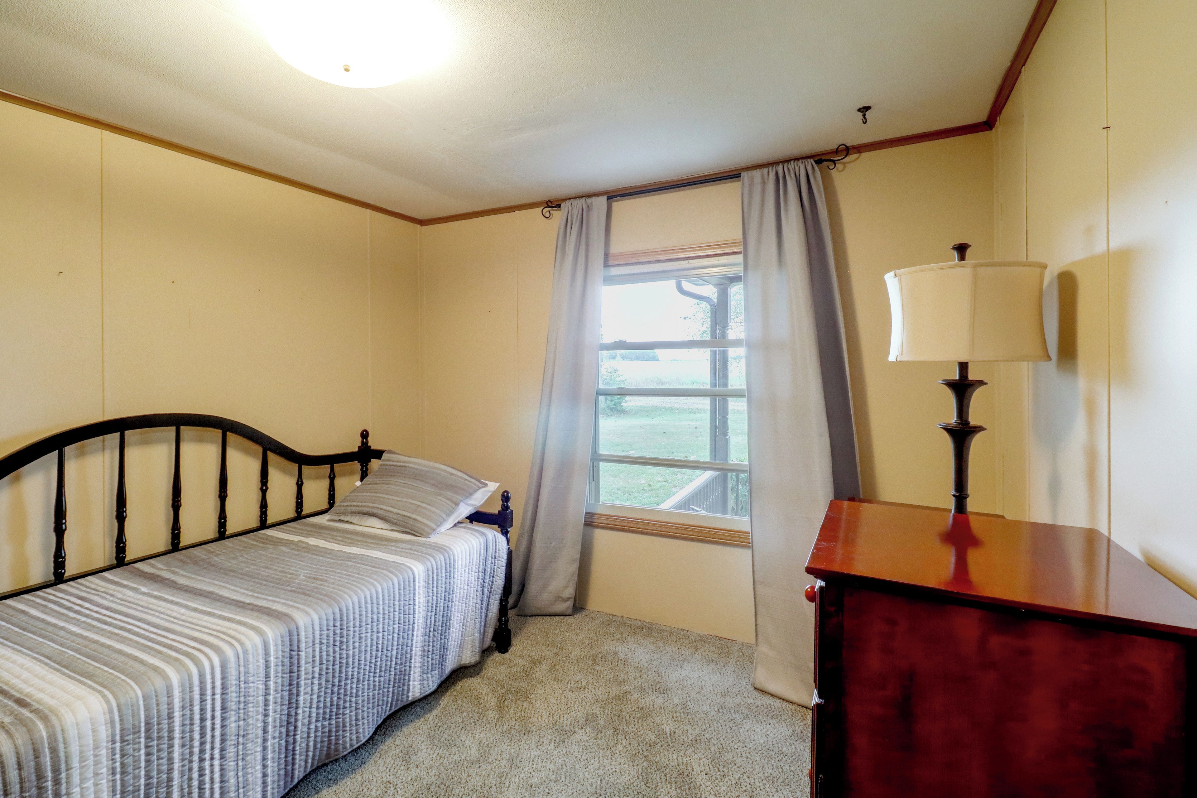 Une petite chambre avec un lit simple, une tête de lit en bois, une table de chevet avec une lampe, une fenêtre avec des rideaux et une vue sur un espace verdoyant.