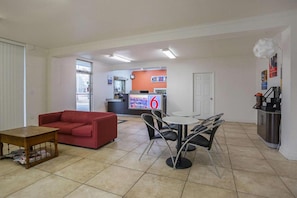 A living room with a red sofa, a coffee table, and a dining area with a table and chairs. There is a kitchen area in the background with a counter and a refrigerator.