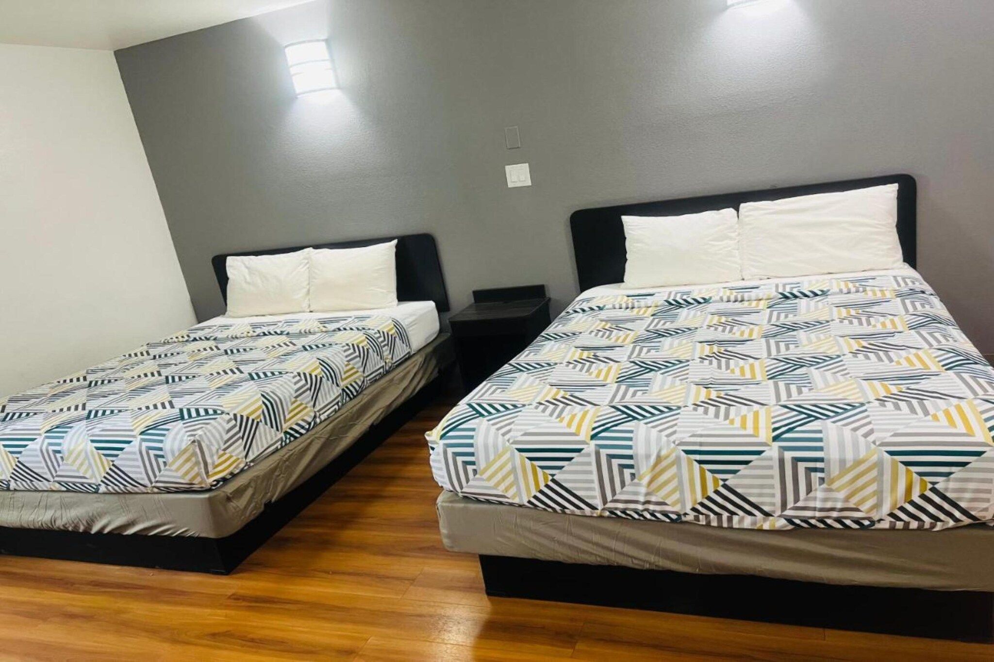 Dos camas individuales con ropa de cama estampada, suelo de madera y pared gris con una lámpara.