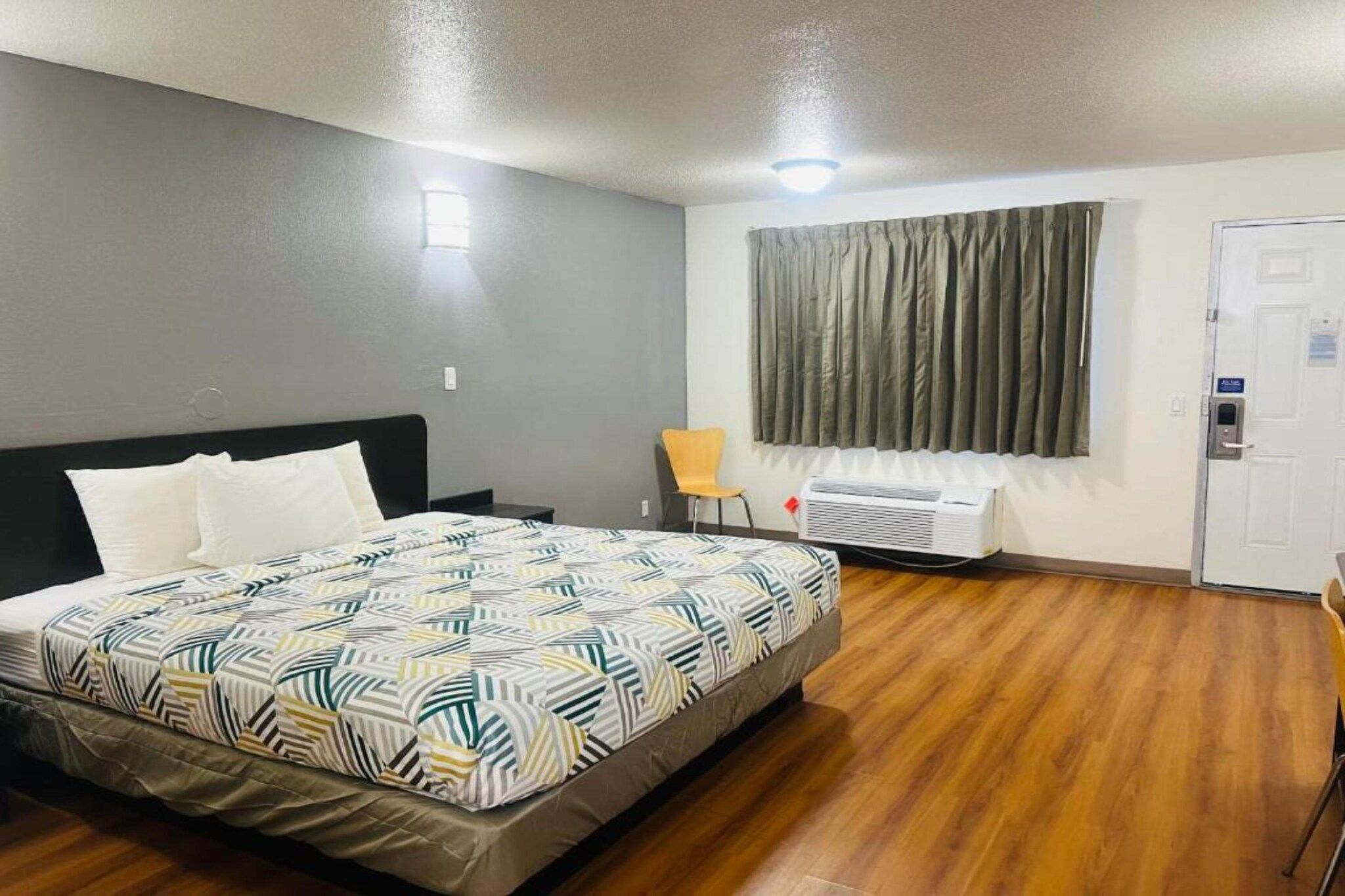 A hotel room with a bed, a chair, a window with curtains, a door, and an air conditioning unit.