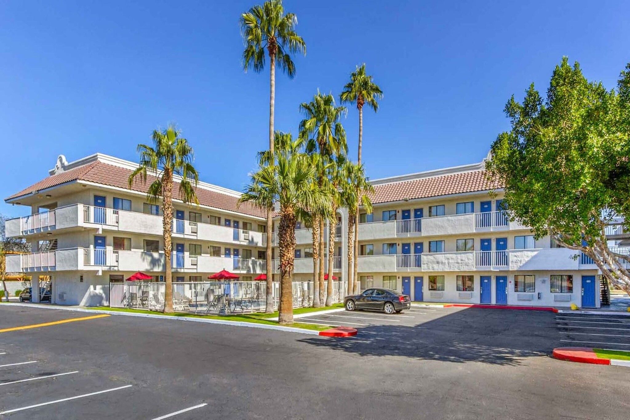 A multi-story motel with a parking lot, palm trees, and a clear blue sky.