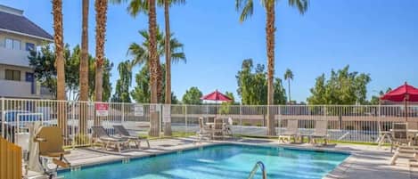 A swimming pool with lounge chairs and umbrellas, surrounded by palm trees and a fence.