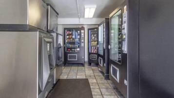 Un couloir étroit, avec des distributeurs automatiques et un réfrigérateur d’un côté, et une armoire de couleur sombre de l’autre.