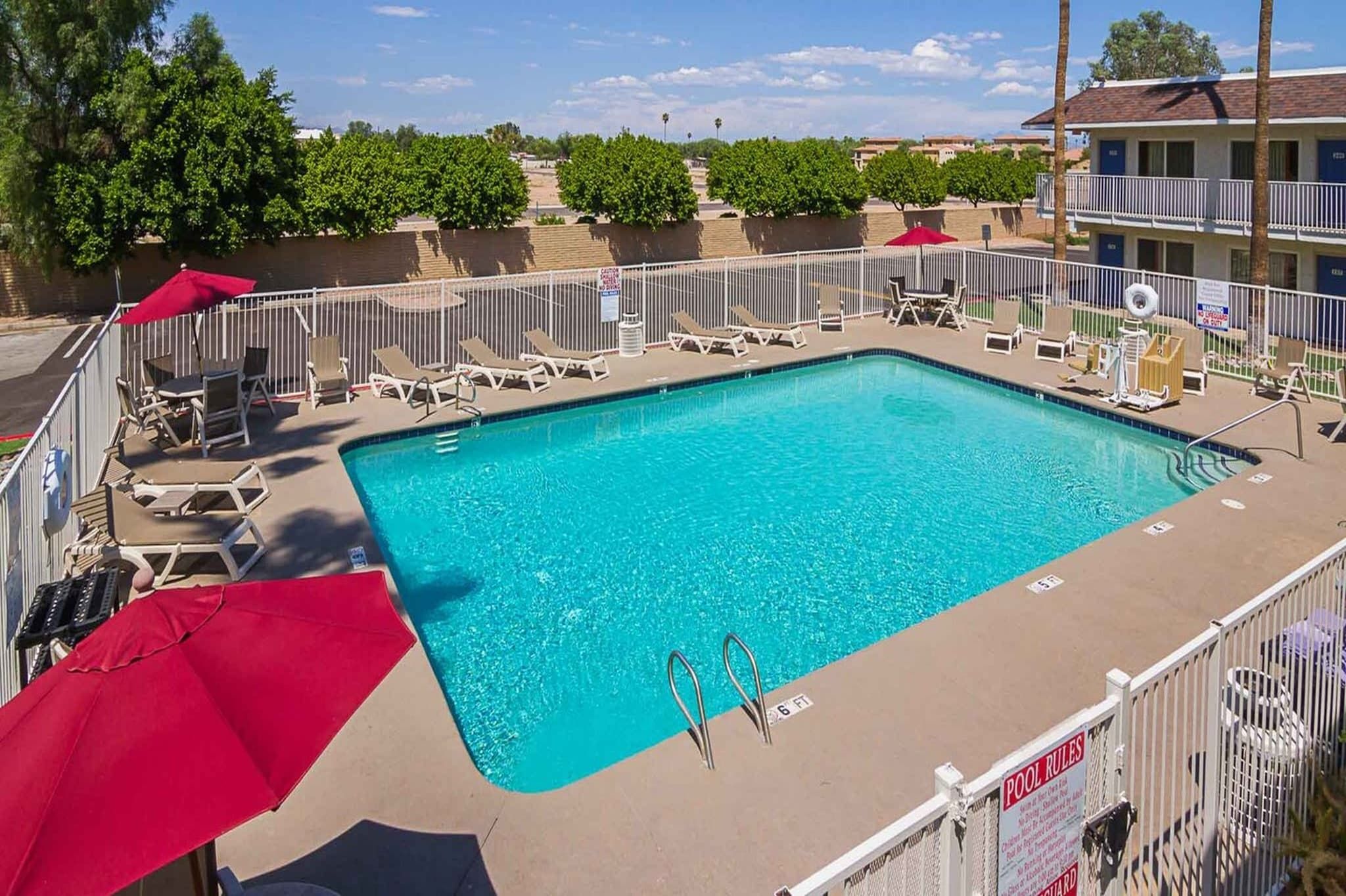 A rectangular swimming pool with a clear blue water surface, surrounded by a white fence. There are lounge chairs and umbrellas along the poolside.