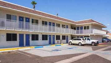 A two-story motel with a parking lot, a white car parked in front, and a palm tree on the roof.