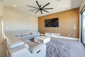 A modern living room with a sectional sofa, a flat-screen TV mounted on a wooden wall, and a ceiling fan. - 1 Mi to South Mtn Park! Home w/ Poolside Oasis (Phoenix)
